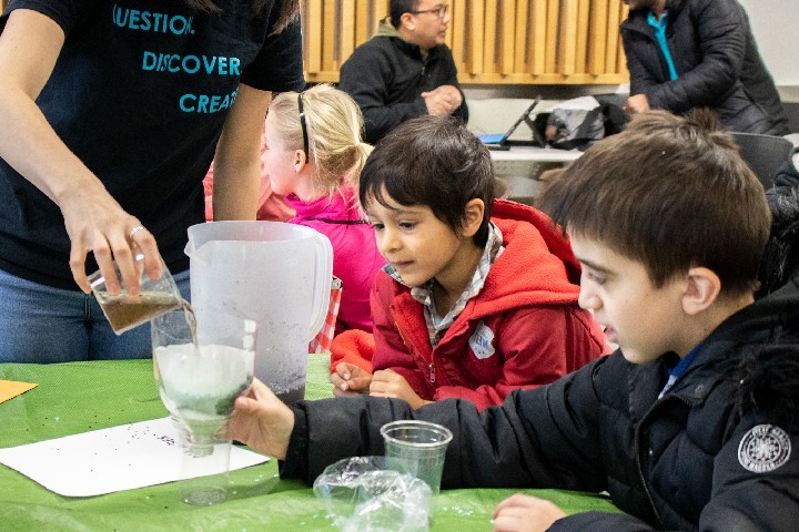 At a MCYU in the City Workshop a MCYU facilitator pours a mixture into a water filter that two boys created. Parents talking in the background.